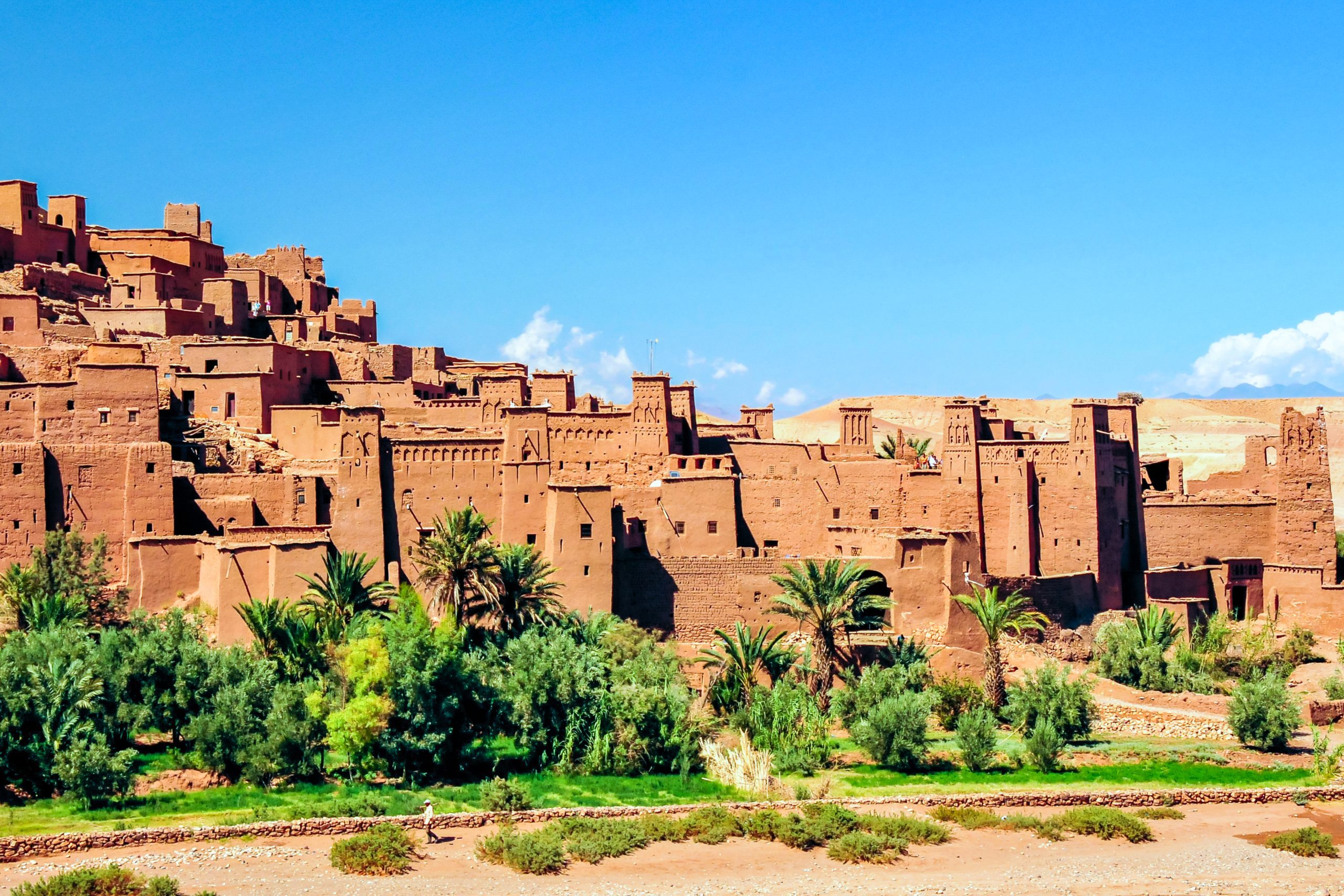 Ancient clay buildings of Kasbah Aït Benhaddou rising from desert landscape, with palm trees and green plants in the foreground under a bright blue sky
