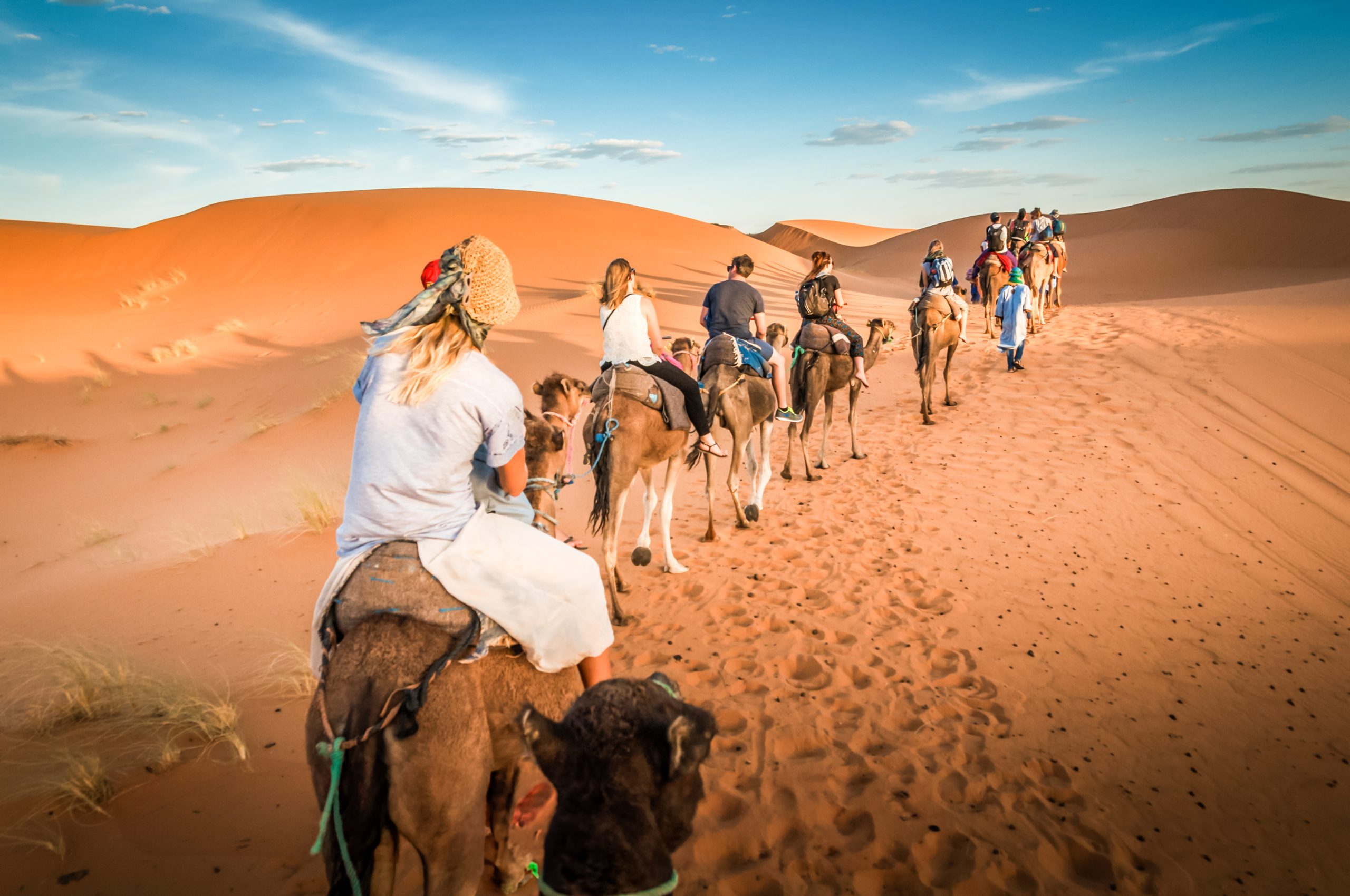 Group of tourists riding camels in a line across orange sand dunes in the desert under a blue sky with white clouds