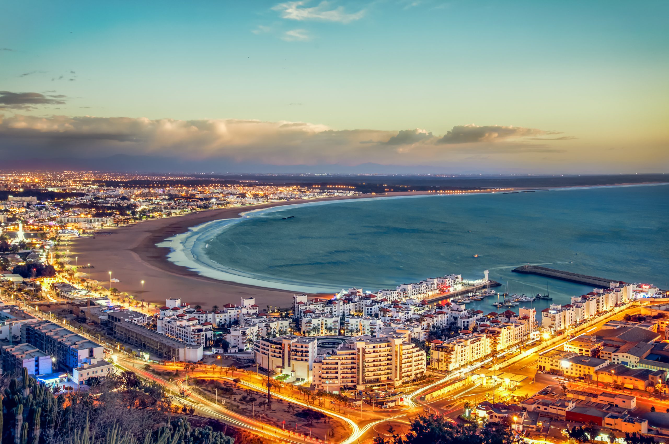 Agadir city at sunset showing curved beach, blue ocean, white buildings, and bright lights along the coastline with mountains in the background