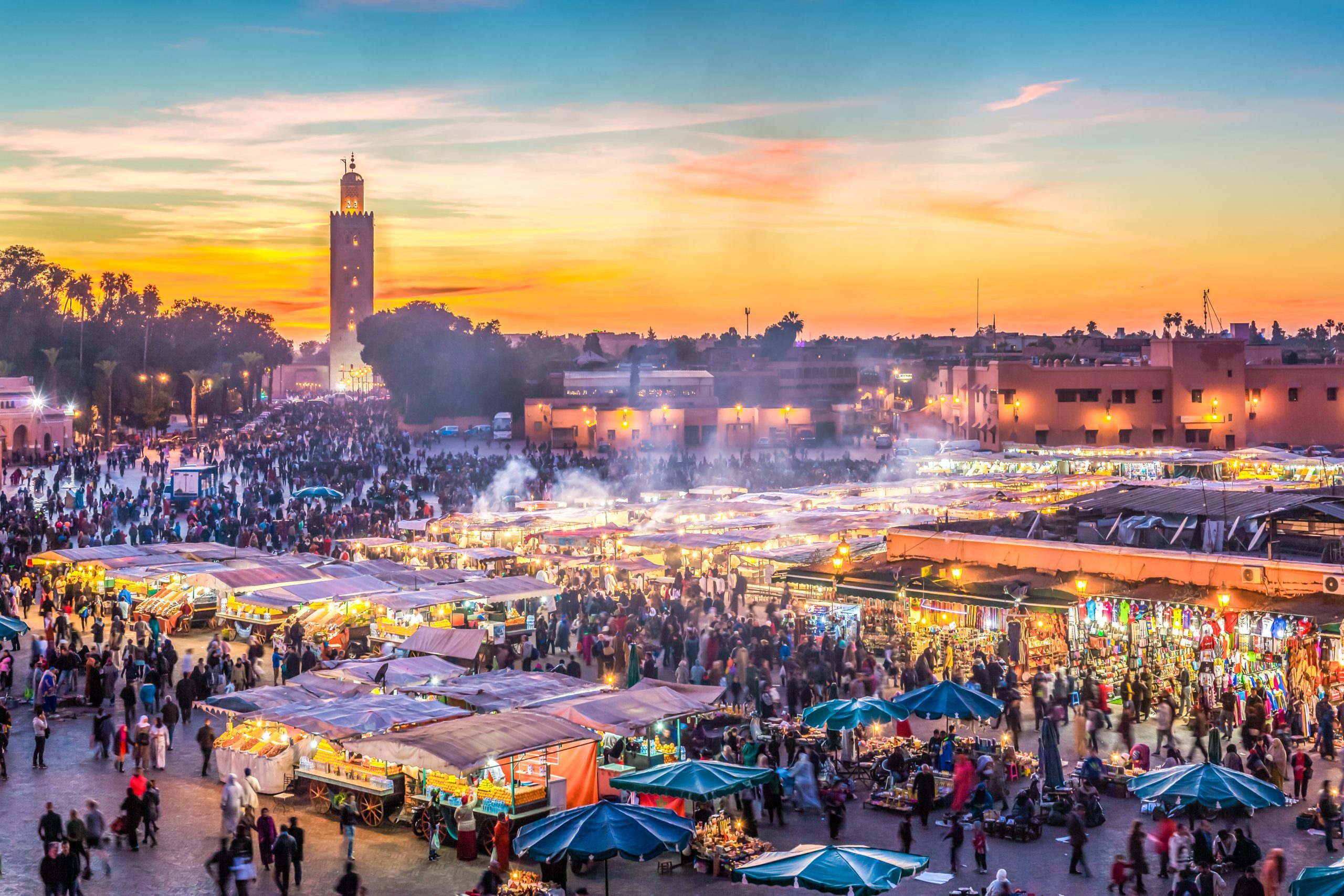 Busy Jemaa el-Fnaa square in Marrakech at sunset with crowds of people, food stalls, and market tents, with Koutoubia mosque tower in the background and colorful sky