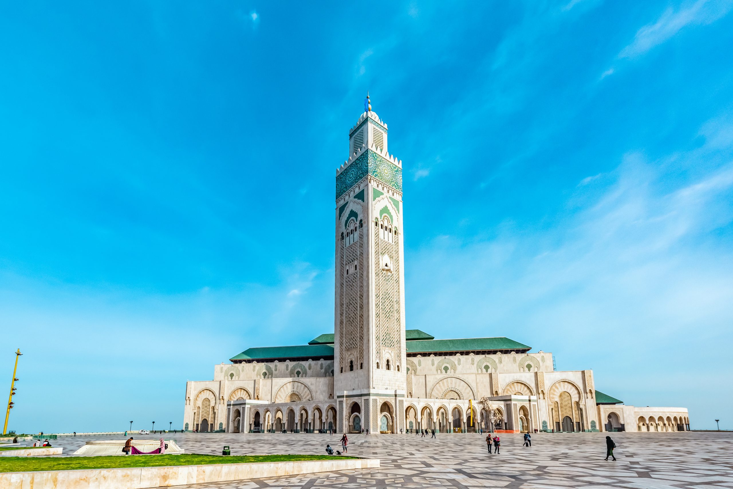 Hassan II Mosque with its tall white tower and green roof against blue sky, with tourists walking on large stone plaza in front