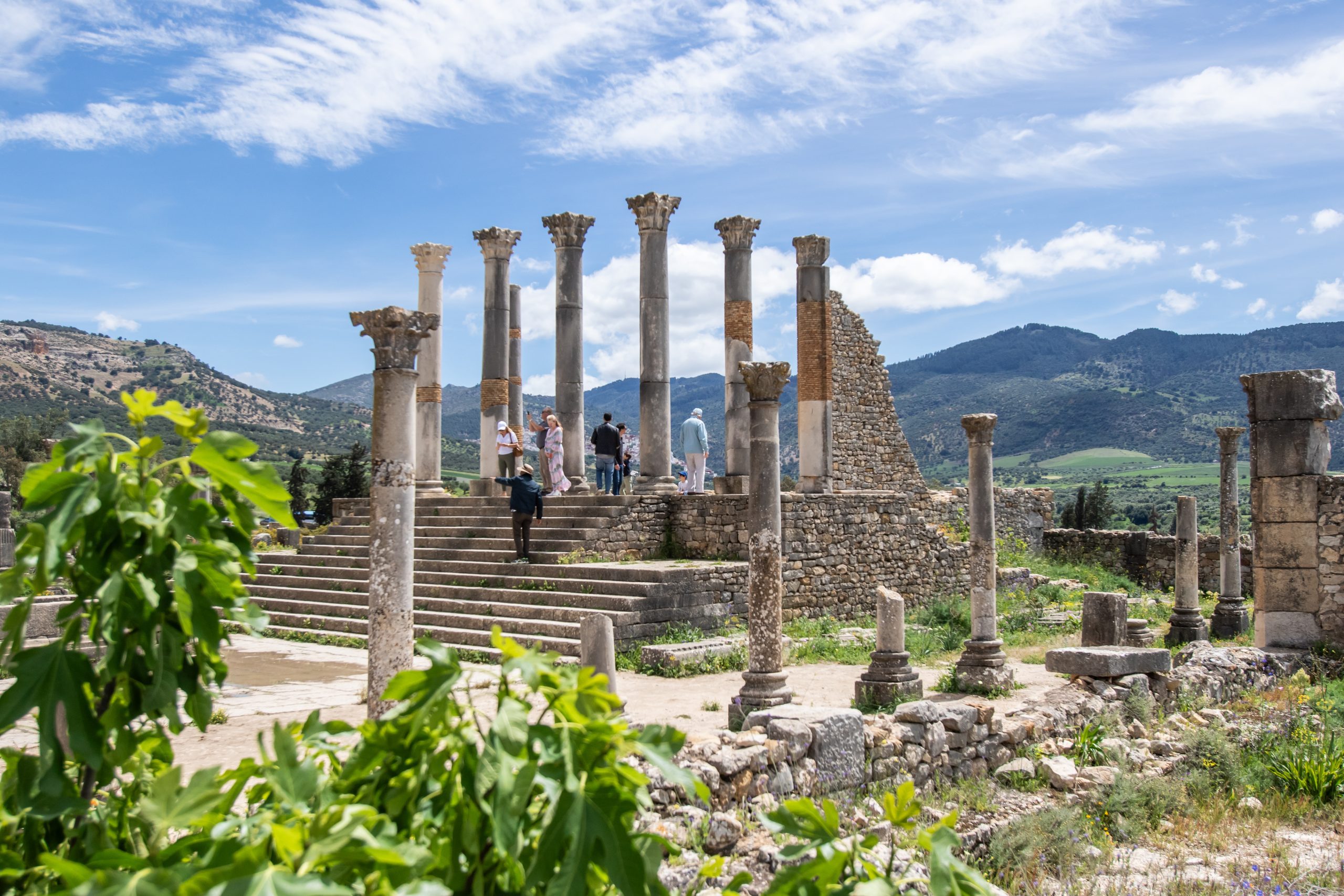 Ancient stone columns and ruins at Volubilis with tourists walking among them and green hills behind