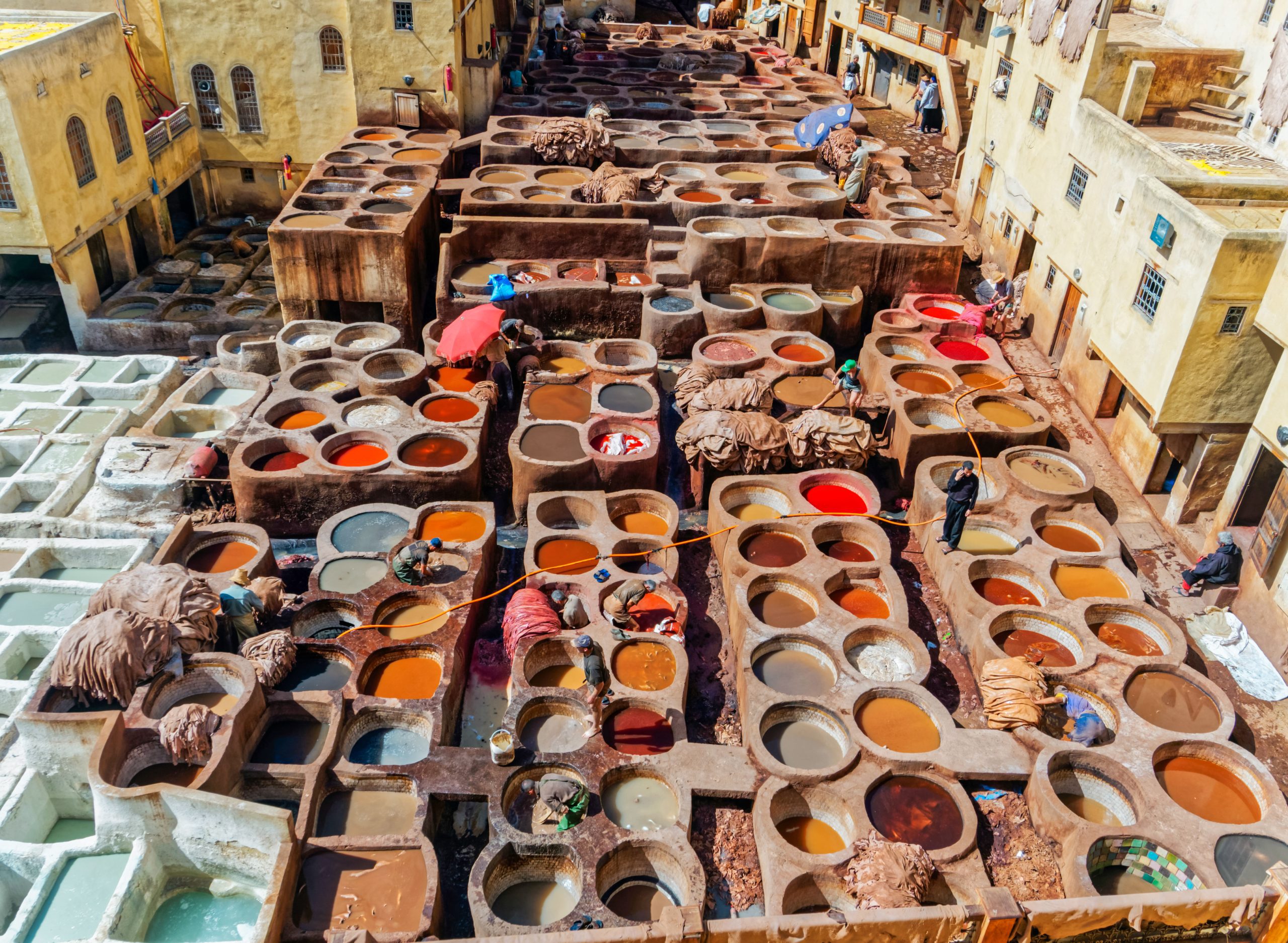 Tannery in Fes showing many round stone pools filled with colorful dyes in red, orange, yellow, and brown, with workers processing leather hides